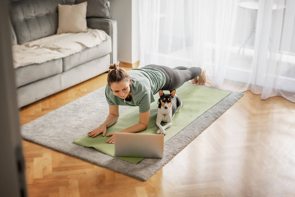 Cheerful beautiful woman practicing yoga at home online on the carpet and yoga mate with her dog nearby, sports mental health - Photo, Image