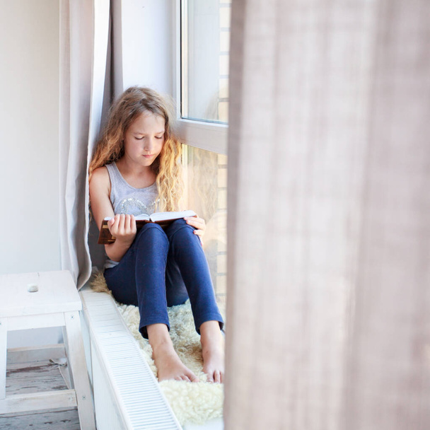 Child reading book at home. Girl sitting at window at read - 写真・画像