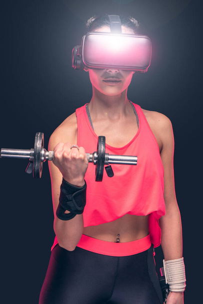 Schöne aktive junge sportliche kaukasische Athletin beim Training in rosafarbenem Unterhemd - Studioshooting mit hübscher schlanker junger sportlicher Brünette beim Training mit einer futuristischen High-Tech-Brille - Foto, Bild