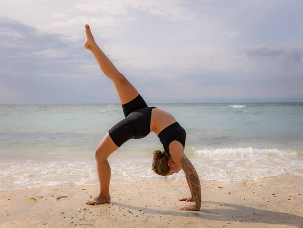 Yoga de praia. Jovem praticando Eka Pada Chakrasana, One Legged Wheel Pose. Posição de proa virada para cima é uma backbend profunda. Flexibilidade, força, paciência. Retiro de ioga. Thomas beach, Bali, Indonésia - Foto, Imagem