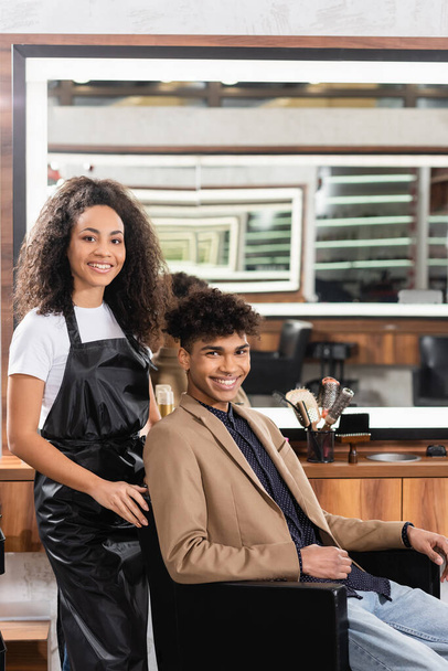 Africano americano cabeleireiro no avental sorrindo perto do cliente na cadeira  - Foto, Imagem