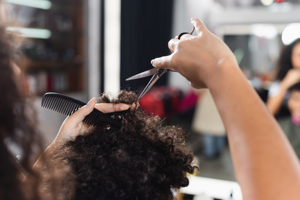Vue recadrée de coiffeur afro-américain sur le premier plan flou tenant peigne et couper les cheveux de l'homme  - Photo, image