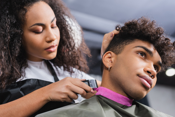 Vista de ángulo bajo del hombre afroamericano sentado cerca de la peluquería que sostiene trimmer sobre fondo borroso  - Foto, imagen