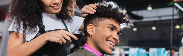 Sonriente hombre afroamericano sentado cerca de peluquería con trimmer en el salón, pancarta  - Foto, imagen