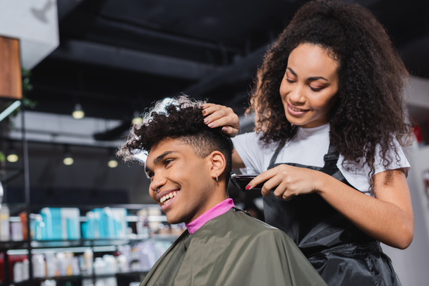 Улыбающийся африканский американский парикмахер в фартуке обрезает шею клиенту в салоне  - Фото, изображение