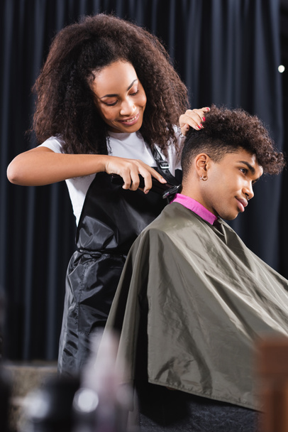 Happy african american hairdresser trimming neck of young man in cape and collar paper - Фото, зображення