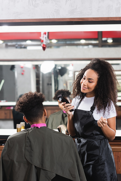 Sorridente parrucchiere africano americano che tiene trimmer e guarda il cliente nel salone  - Foto, immagini