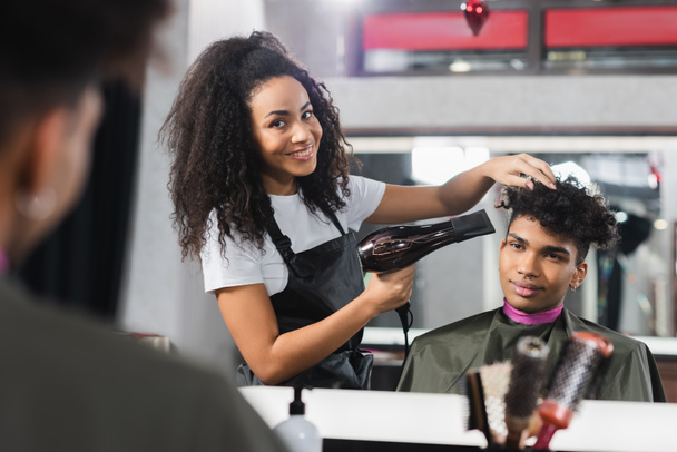 Cheerful african american hairstylist drying hair of young client on blurred foreground  - Photo, Image