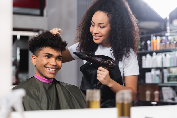 Glimlachende klant op zoek naar spiegel in de buurt van hairstylist met haardroger in salon  - Foto, afbeelding