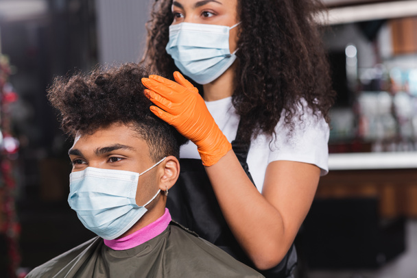 cliente afroamericano en máscara médica sentado cerca de peluquero sobre fondo borroso  - Foto, imagen
