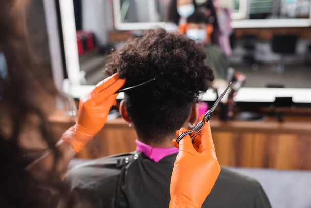 Ножиці і гребінець в руках африканського американського перукаря в рукавичках латексу поруч з клієнтом.  - Фото, зображення