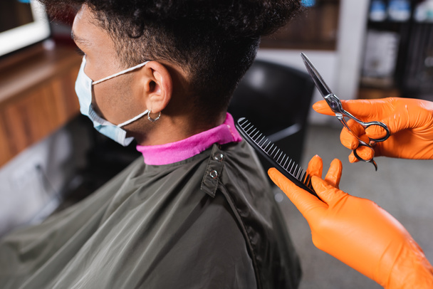 Перукар в латексних рукавичках тримає гребінець і ножиці біля афроамериканського клієнта в медичній масці
  - Фото, зображення