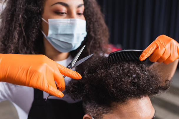 Scissors and comb in hands of african american hairstylist working with client in salon during quarantine  - 写真・画像