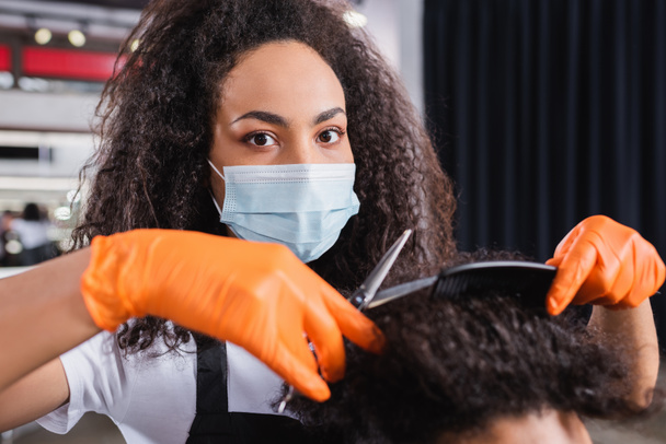 African american hairstylist in latex gloves and medical mask holding scissors and comb near hair of blurred man  - Photo, Image