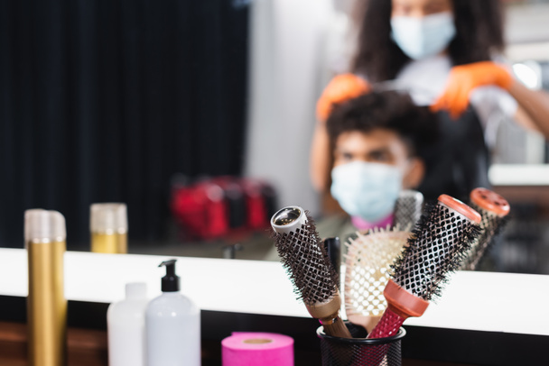 Cepillos cerca de espejo y peluquería afroamericana con cliente sobre fondo borroso en el salón  - Foto, Imagen