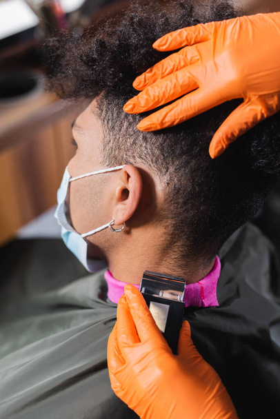 Hairstylist in latex handschoenen trimmen nek van Afrikaanse Amerikaanse man in medisch masker en cape  - Foto, afbeelding