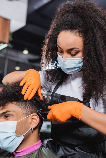 African american hairdresser in medical mask and gloves trimming hair of client on blurred foreground  - Fotografie, Obrázek