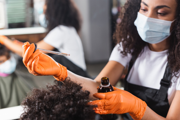 Olej kosmetyczny w rękach afrykańskiego fryzjera w lateksowych rękawiczkach i klienta na nieostrym pierwszym planie  - Zdjęcie, obraz