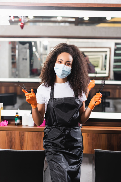 African american hairdresser in medical mask holding comb and scissors  - Foto, Bild