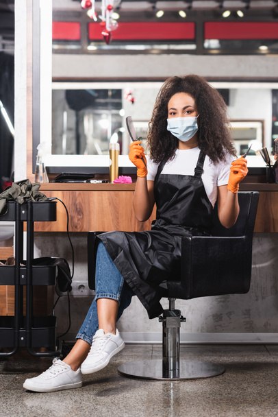 coiffeur afro-américain en masque médical tenant des ciseaux et peigne dans la chaise  - Photo, image