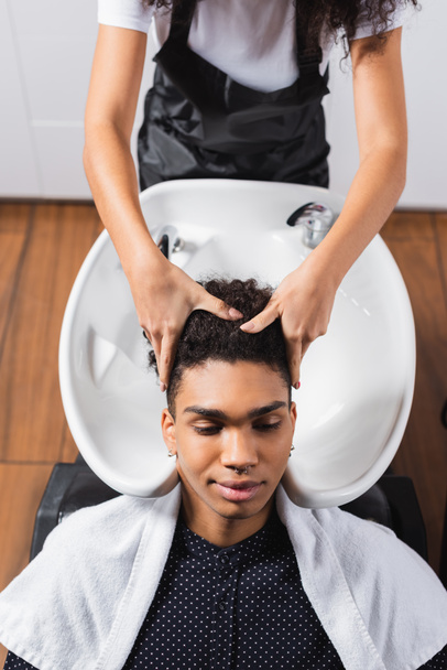 Vue grand angle du client afro-américain en serviette assis près du coiffeur et évier sur fond flou  - Photo, image
