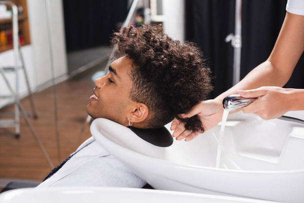 Sonriente cliente afroamericano sentado cerca de peluquería con ducha  - Foto, Imagen