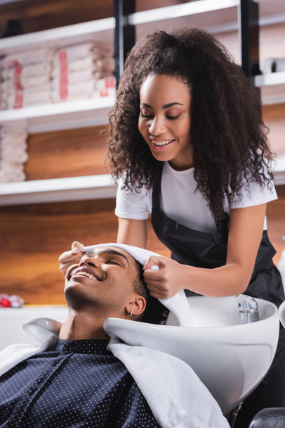 Coiffeur afro-américain souriant tenant une serviette près de la tête du client dans le salon  - Photo, image