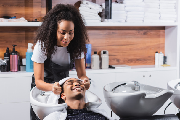Cheerful african american man sitting near sink and hairdresser with towel in salon  - 写真・画像
