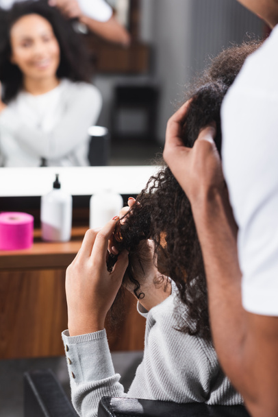 African american hairdresser on blurred foreground touching curly hair of client  - Photo, Image