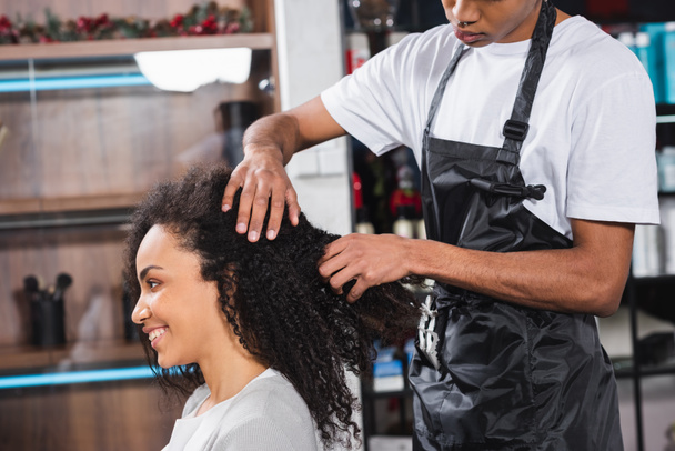 Uśmiechnięta Afroamerykanka siedząca obok fryzjera w fartuchu  - Zdjęcie, obraz