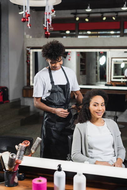 Smiling woman sitting near african american hairdresser with hair iron  - Zdjęcie, obraz