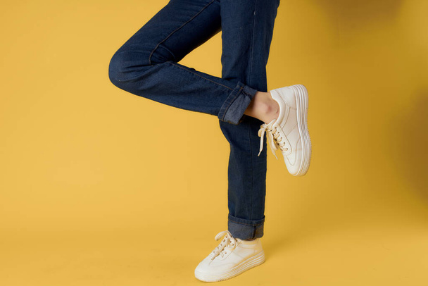 voet gebaar witte sneakers mode jeans straat stijl gele achtergrond - Foto, afbeelding