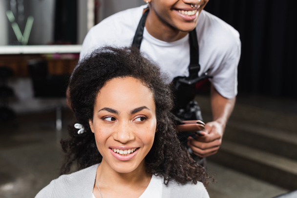 Mujer afroamericana sonriente mirando hacia otro lado cerca de un peluquero borroso con plancha para el cabello  - Foto, imagen