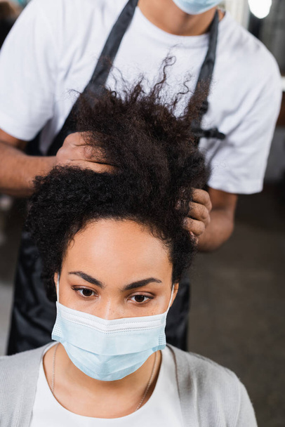 Femme afro-américaine en masque médical assis près du coiffeur sur fond flou  - Photo, image