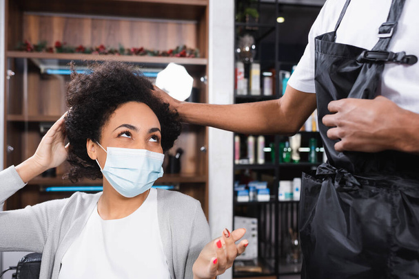 African american woman in medical mask looking at hairdresser in salon  - Fotó, kép