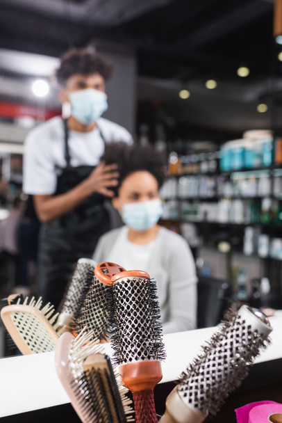 Escovas de cabelo perto de espelho e cliente afro-americano borrado em máscara médica e cabeleireiro  - Foto, Imagem