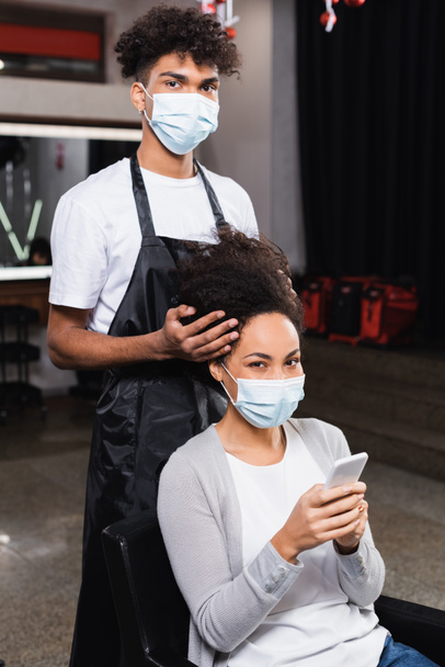 African american woman with smartphone looking at camera near hairstylist in medical mask  - Φωτογραφία, εικόνα