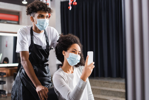 African american woman in medical mask holding smartphone near hairdresser in salon  - Фото, изображение