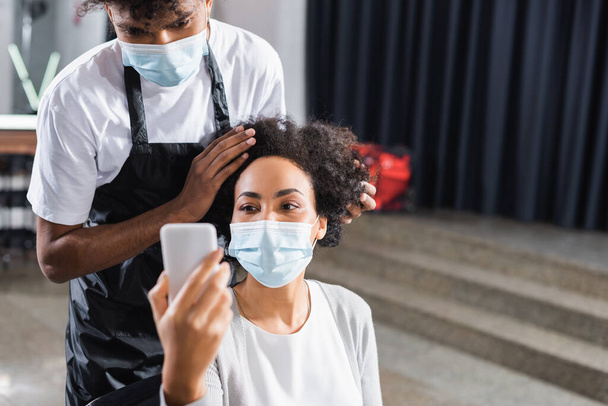 African american client in medical mask using smartphone on blurred foreground near hairstylist  - Foto, Bild