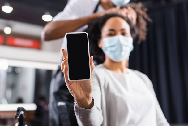 Smartphone in hand of african american woman in medical mask near hairstylist on blurred background  - Fotoğraf, Görsel