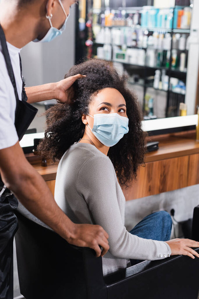 African american woman in medical mask looking at blurred hairstylist  - Foto, afbeelding