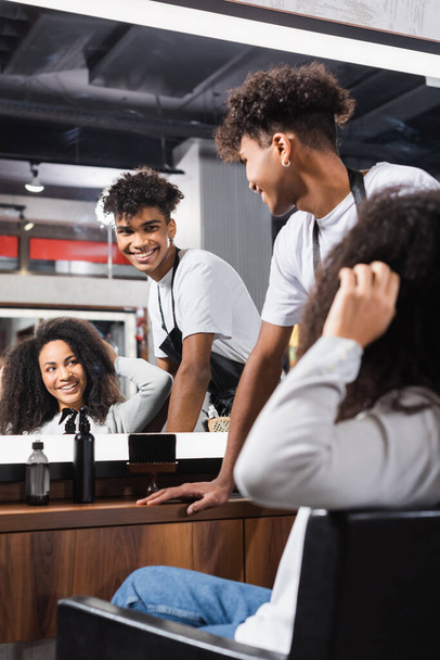 Acconciatore afroamericano sorridente che guarda lo specchio vicino alla donna che tocca i capelli in primo piano sfocato  - Foto, immagini