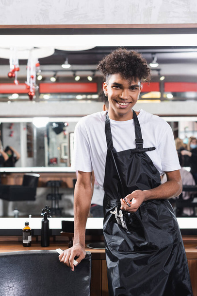 Smiling african american hairdresser in apron holding comb and scissors  - Foto, Bild