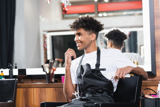 Jeune coiffeur afro-américain souriant tout en tenant peigne et ciseaux dans le salon  - Photo, image