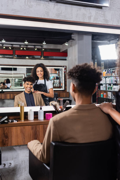 Улыбающийся африканский американский парикмахер обнимает клиента в салоне  - Фото, изображение