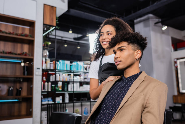 Positive african american hairstylist on blurred background standing near young man  - Foto, afbeelding