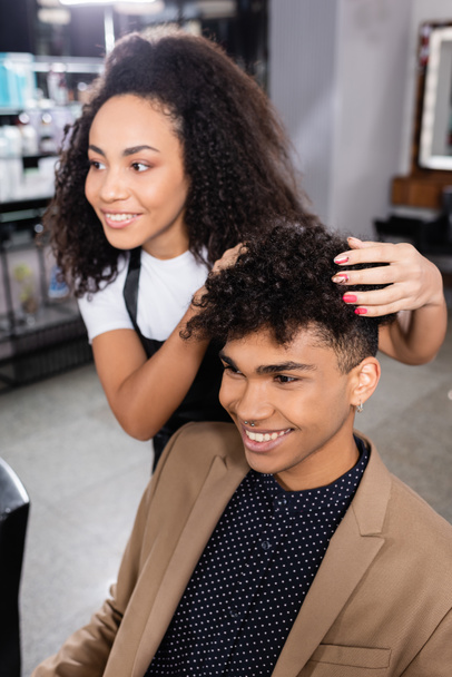 Positive man sitting near blurred african american hairdresser touching hair  - Фото, зображення
