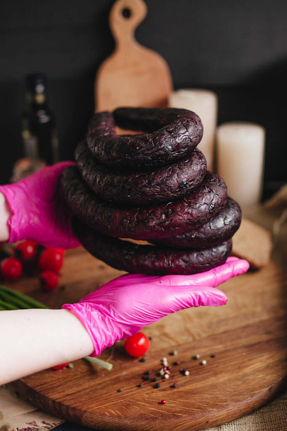 Person in pink gloves holds blood sausage and cherry tomatoes in the background on a wooden board in the kitchen. - Photo, Image