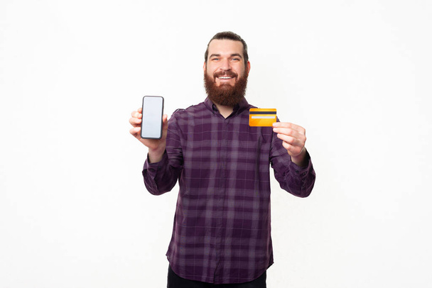 Vrolijke knappe man met baard in geblokt shirt met smartphone en creditcard. - Foto, afbeelding