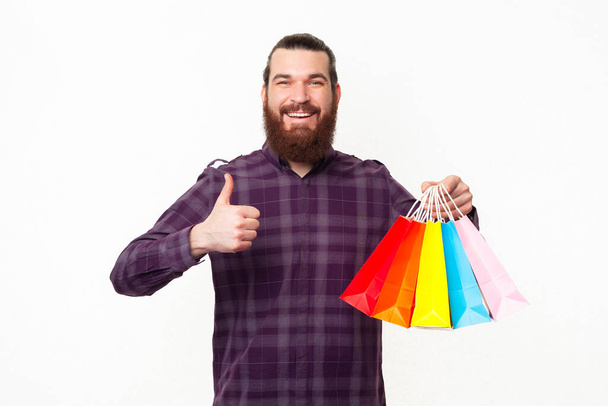 Kareli tişörtlü mutlu, sakallı adam başparmağını kaldırıyor ve renkli alışveriş torbaları tutuyor.. - Fotoğraf, Görsel